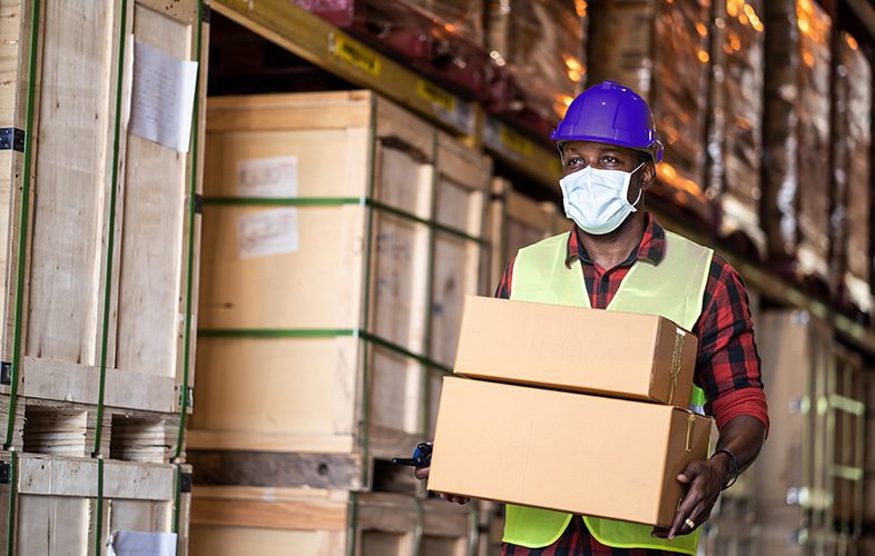 Imagem de um funcionário com seus equipamentos de seguranças, segurando duas caixas de papelão. Ao fundo, há caixas empilhadas com produtos importados, simbolizando a nacionalização de produtos.