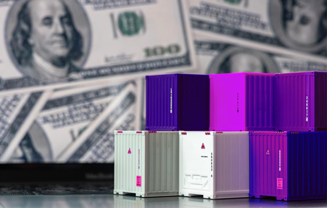 Containers roxos em cima de notebook com dólares na tela, simbolizando os impostos de importação.