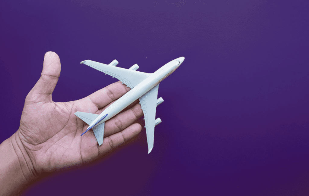 Avião em miniatura com branca, detalhes em azul sobre uma mão com a palma aberta, em fundo roxo, simbolizando o modal de transporte.